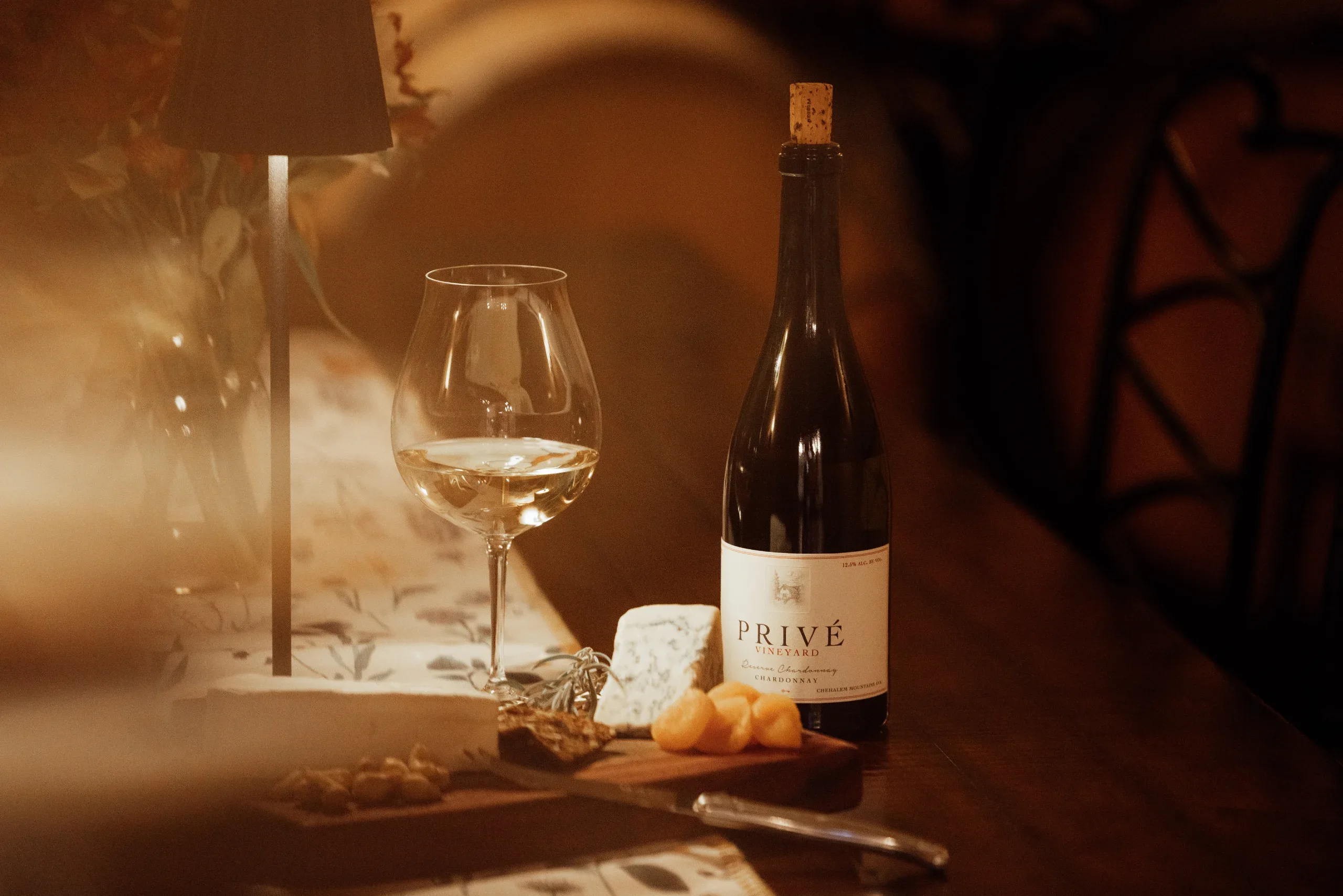 A glass of white wine beside a bottle of Privé and a cheese board